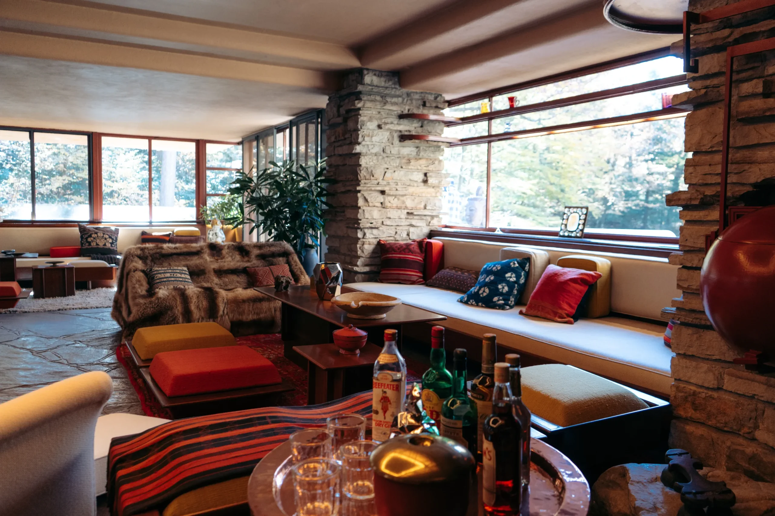 Salon w domu Fallingwater autorstwa Franka Lloyda Wrighta z widokiem na otoczenie natury, wyposażony w prostą kanapę, pufy i dywan.