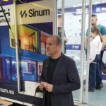 Przemysław Sobczyk, Szef Sprzedaży w Tech Sterowniki, na stoisku firmy podczas targów nieruchomości w hali Urania w Olsztynie 2025. W tle ekspozycja systemu Sinum oraz inteligentnych sterowników ogrzewania i oświetlenia.
